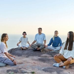 Five adults meditating outdoors on a mountain top during sunrise, promoting relaxation and mindfulness.