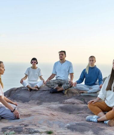Five adults meditating outdoors on a mountain top during sunrise, promoting relaxation and mindfulness.