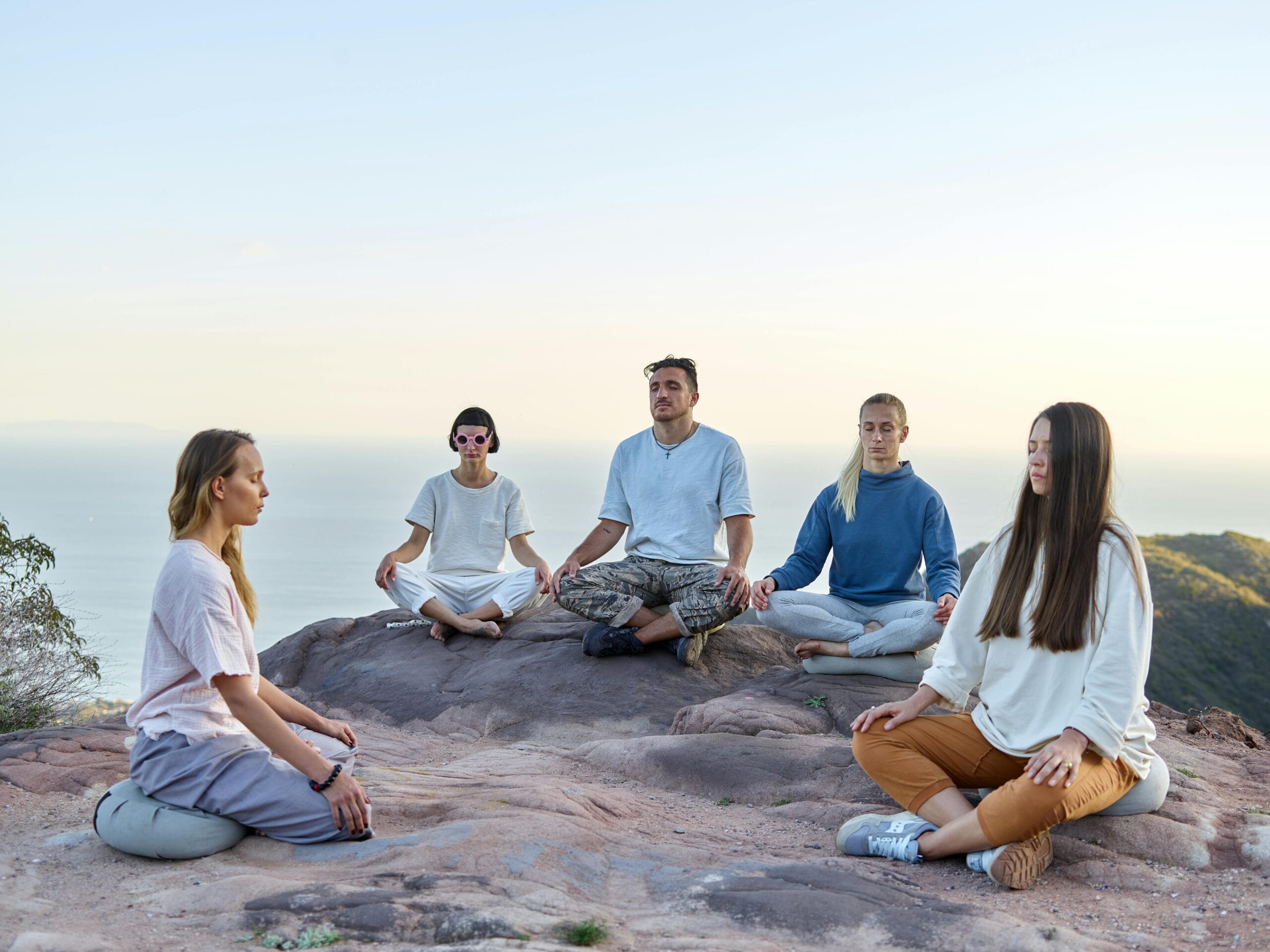 Five adults meditating outdoors on a mountain top during sunrise, promoting relaxation and mindfulness.